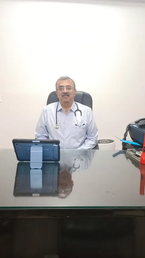 Dr Deepak Ahuja seated at his consulting desk, wearing a stethoscope around his neck, with a tablet and a telephone on the desk in front of him.